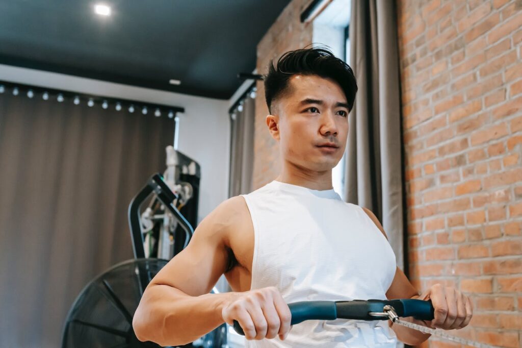 A man of asian origin using his rowing machine, with his exercise bike from his home gym equipment list visible in the background
