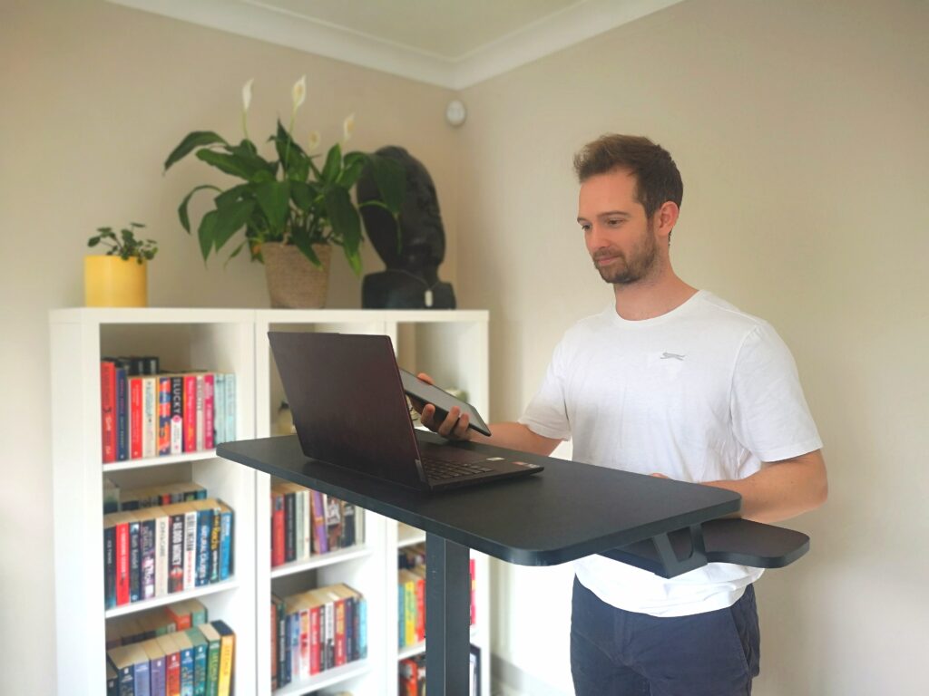 A photo of a male online personal trainer for weight loss standing at his desk with a laptop, speaking to a client via video call