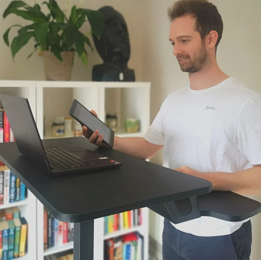 A photo of a male online personal trainer for weight loss standing at his desk with a laptop, speaking to a client via video call